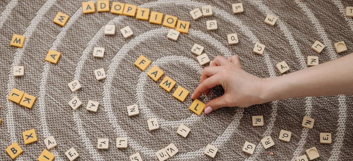 A hand arranges letter tiles spelling 'family' and 'adoption' on a circular patterned carpet.